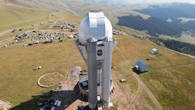 The Assy-Turgen Observatory is high in the mountains. There is a tent camp next to the observatory. Large cumulus clouds in a blue sky. Yellow-green hills, forest in places. Top view from a drone