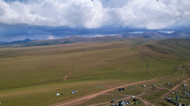 Big white clouds over green hills and mountains. Top view from the drone on endless fields. Roads are visible in places, herds of animals graze. A tent camp has been set up. Coniferous trees in gorge