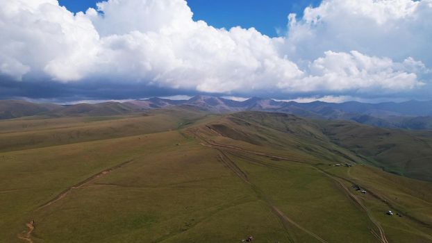 Big white clouds over green hills and mountains. Top view from the drone on endless fields. Roads are visible in places, herds of animals graze. A tent camp has been set up. Coniferous trees in gorge