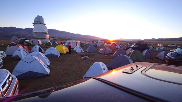 Sunrise over the camp near the Assi Observatory. The sun comes out from behind the high hills. Red-yellow color. Lots of tents and cars. The guy launches the drone. Telescopes are covered with domes