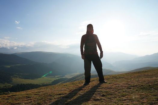 A guy on the edge of the hill admires the view. In the distance you can see high mountains and green hills covered with forest. Wide margins. Clouds are visible and sun is shining brightly