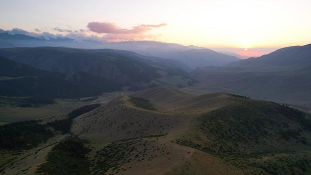 Red rays of the sun at sunset among the mountains. Green grass turns yellow, in places a small forest. Tourists walk in the hills. A river runs in the distance. The clouds are highlighted in pink.