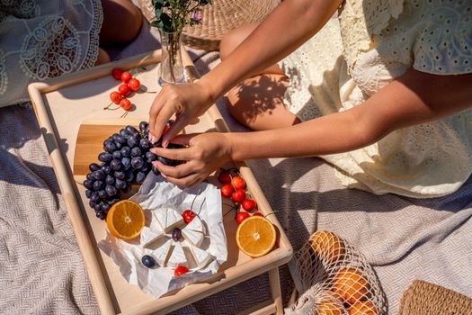 Romantic picnic for two with fruit, bread and cheese. Oranges, cherries, black grapes and camembert on a wooden table. The girl's hands with a manicure tear off a grape berry sitting on a light blanket