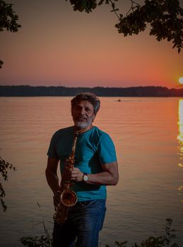 An adult man plays the saxophone at sunset by the river in the evening.