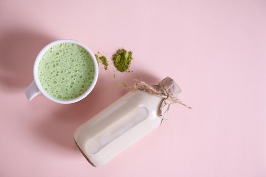 Still life. Bottle of organic plant based milk on pink surface near white ceramic cup of antioxidant healthy drink- whipped matcha latte with foam and a heap of powdered japanese green tea. Copy space