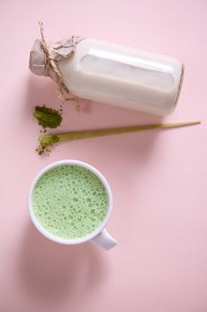 Flat lay. Vertical studio shot of a white ceramic cup of whipped matcha latte with foam, bamboo scoop with powdered green tea, bottle of healthy organic wholesome raw vegan milk over pink background