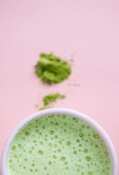 Selective focus on a white cup of whipped green tea with foam and blurred powdered japanese matcha, over pink background with copy advertising space. Antioxidant healthy drink. Oriental traditions