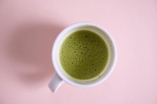 High angle view of a white ceramic cup with whipped matcha japanese green tea, shot in studio with soft shadow, isolated on pink background with copy advertising space. Flat lay. Still life