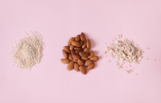 Horizontal studio shot of healthy raw organic almond nuts, sesame seeds and oat-flakes scattered on pink background. Ingredients for making plant based milk. Concept of Vegan, Veganism. Healthy eating