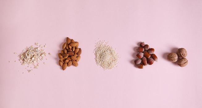 Flat lay stacks of organic raw vegan superfood: sesame seeds, almond nuts, oat-flakes, walnut and hazelnut scattered on pink background, copy space. Horizontal studio shot. Still life. Selective focus