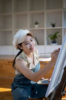 Asian woman painter sitting in front of the canvas and painting with water color at workshop.