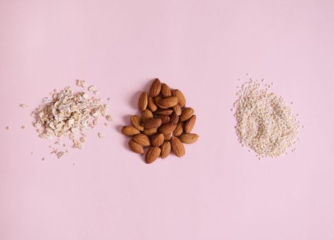 Flat lay composition with stacks of organic raw vegan superfood: almonds, sesame seeds and oat-flakes, scattered on pink background with copy space for advertising text. Veganism and healthy eating