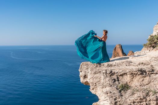 A girl with loose hair in a long mint dress descends the stairs between the yellow rocks overlooking the sea. A rock can be seen in the sea. Sunny path on the sea from the rising sun.