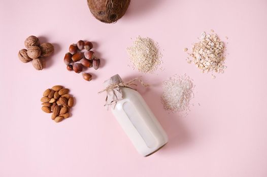 Flat lay. Still life with organic plant based milk in a glass bottle and healthy nuts, cereals and seeds: almonds, hazelnuts, walnuts, rice, oatmeal, sesame and coconut on pink background. Copy space