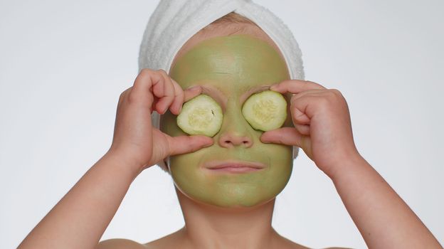 Smiling child girl applying cleansing moisturizing green facial mask, holding slices of cucumber, covering eyes. Teenager kid face skin care treatment natural cosmetics. Female close-up macro portrait
