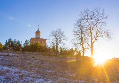 Orthodox church on a hill near the forest. Winter sunset landscape with clear blue sky and sun rays.