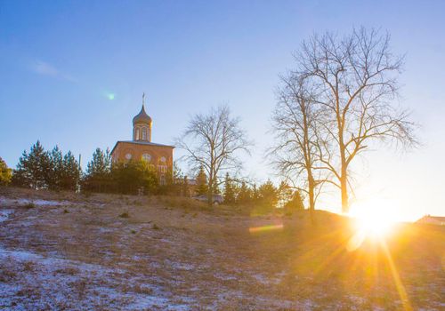 Orthodox church on a hill near the forest. Winter sunset landscape with clear blue sky and sun rays.