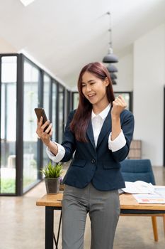 Excited asian business woman waving fists, got good news on smart phone while working at modern office