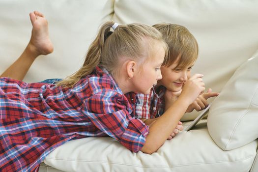 family, children, technology and home concept - smiling brother and sister with tablet pc computer on the sofa in living room