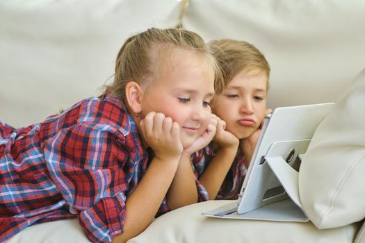 family, children, technology and home concept - smiling brother and sister with tablet pc computer on the sofa in living room