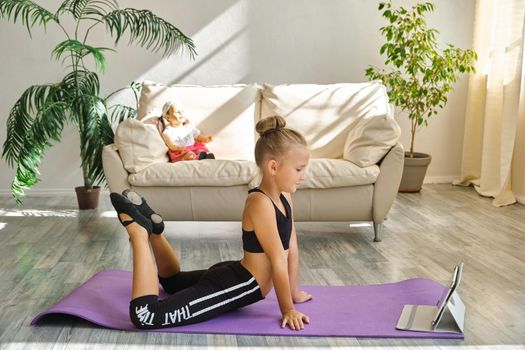 Little cute girl is practicing gymnastics at home. Video lesson. Kid repeating exercises while watching online yoga gymnastics session. Beautiful girl doing fitness exercise at home