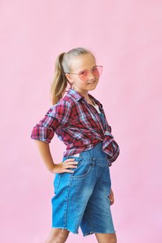 Pretty cool kid girl in pink sunglasses posing against pink background. Fashion style portrait of young happy smiling woman, copy space.