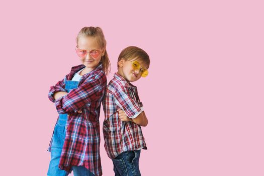 Cute stylish little girl and boy in color sunglasses dressed in plaid shirts, looking at camera and laughing while standing on pink background with copy space.