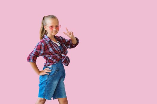 Pretty cool kid girl in pink sunglasses posing against pink background. Fashion style portrait of young happy smiling woman, copy space.