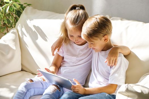 family, children, technology and home concept - smiling brother and sister with tablet pc computer on the sofa in living room