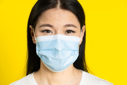 Happy, excited mature asian woman wearing medical face mask coronavirus or monkeypox prevention. Charming middle age woman in white t-shirt and medical mask on yellow background.