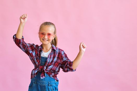 Pretty cool kid girl in pink sunglasses posing against pink background. Fashion style portrait of young happy smiling woman, copy space.