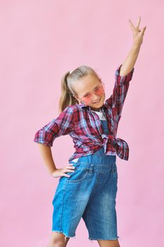 Pretty cool kid girl in pink sunglasses posing against pink background. Fashion style portrait of young happy smiling woman, copy space.