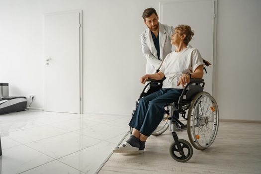 Professional male doctor carrying patient on wheelchair in medicine clinic hall. High quality photo