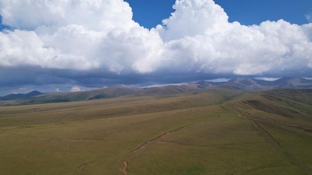 Big white clouds over green hills and mountains. Top view from the drone on endless fields. Roads are visible in places, herds of animals graze. A tent camp has been set up. Coniferous trees in gorge