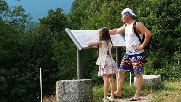 beautiful park in alpine mountains. man and a girl, kid, tourists, study map of park, against backdrop of picturesque landscape of the Alpine mountains and a small town. summer hot day. High quality photo