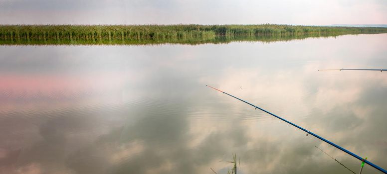 Two fishing rods reflected in the water surface, the reflection of clouds. Quiet hobby, banner.