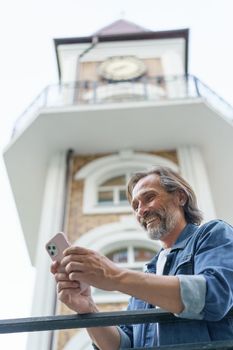 Mature happy grey bearded man laughing reading text message or funny social media looking at smartphone standing in old town wearing casual. Business and travel concept.