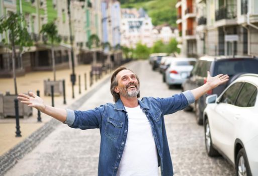 Giving thanks to God mature man standing outdoors with hands spread wide open wearing jeans shirt with white t-shirt under. Mid Aged man glad that life is beautiful standing in old town street.