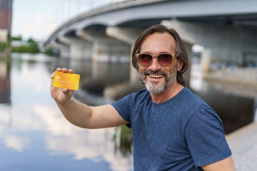 Showing credit, debit card excited freelancer man sitting outdoors next to river working using digital tablet pc, shopping online or paying online banking. E-banking concept. Financial concept.