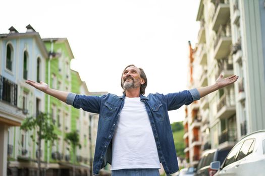 Enjoying life happy mature man spread his hands wide standing on the street of old town wearing white denim shirt. Happy mature man looking sideways sending love outdoor on a summer day.