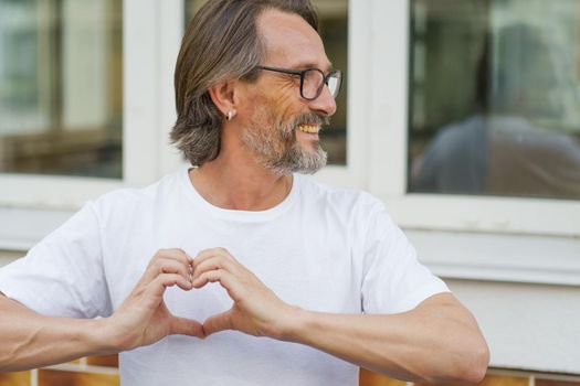 Showing love, gesturing heart happy mature man in glasses smile standing outdoors wearing white t-shirt. Happy mature man in eye glasses and looking sideways sending love outdoor on a summer day.