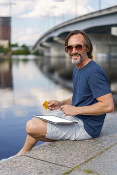 Confident handsome freelancer man hold credit, debit card sitting outdoors next to river working using digital tablet pc, shopping online, paying online banking. E-banking concept. Financial concept.