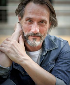 Mature man sitting in city background smiling looking at camera with sexy eye contact . Handsome middle aged grey bearded man smiling on camera old town wearing casual. Business and travel concept.