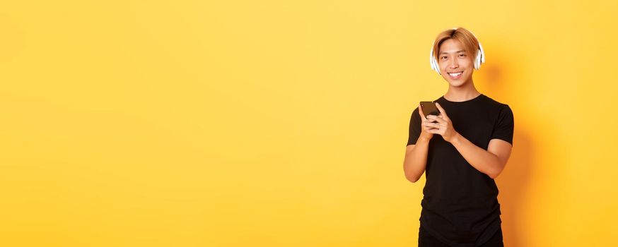 Good-looking smiling, pleased asian guy in wireless headphones, holding smartphone and looking satisfied camera, listening music or podcast.