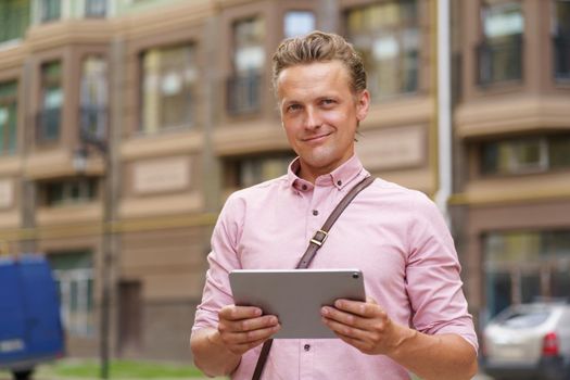 Handsome young man with digital tablet in hands stand outdoors in front urban office building wearing casual pink shirt and shoulder bag. Young freelancer man working on the go.