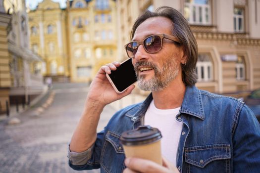 Talking on the phone middle aged bearded man enjoy his take away coffee in paper cup standing outdoors in old town wearing jeans shirt. Freelancer traveling man. Business on the go.