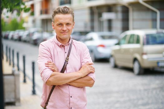 Young handsome man standing on urban streets with arms folded wearing pink shirt, shoulder bag. Happy freelancer business man on old town streets. Travel concept.