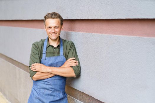 Young handsome employee man in apron take a break standing outdoors leaned on a wall enjoying fresh air after long hard day. Freelancer man at coffee brake enjoying free time.