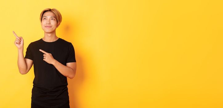 Thoughtful and pleased handsome asian man smiling and pointing fingers upper left corner, standing over yellow background.