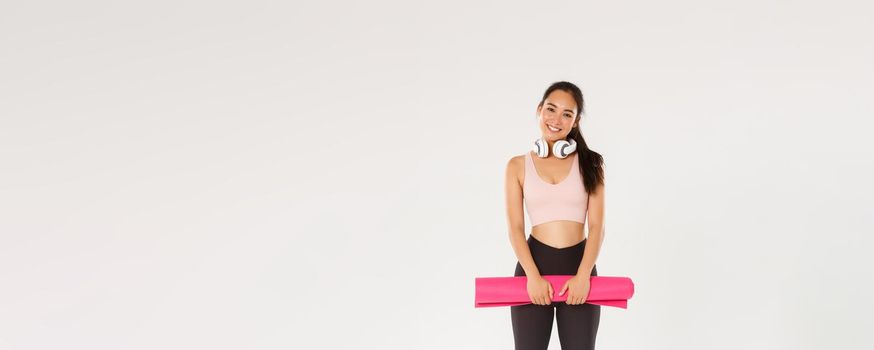 Full length of cute asian girl like fitness, holding rubber mat for exercises or yoga classes, standing in sportswear looking happy after productive training in gym, good workout, white background.